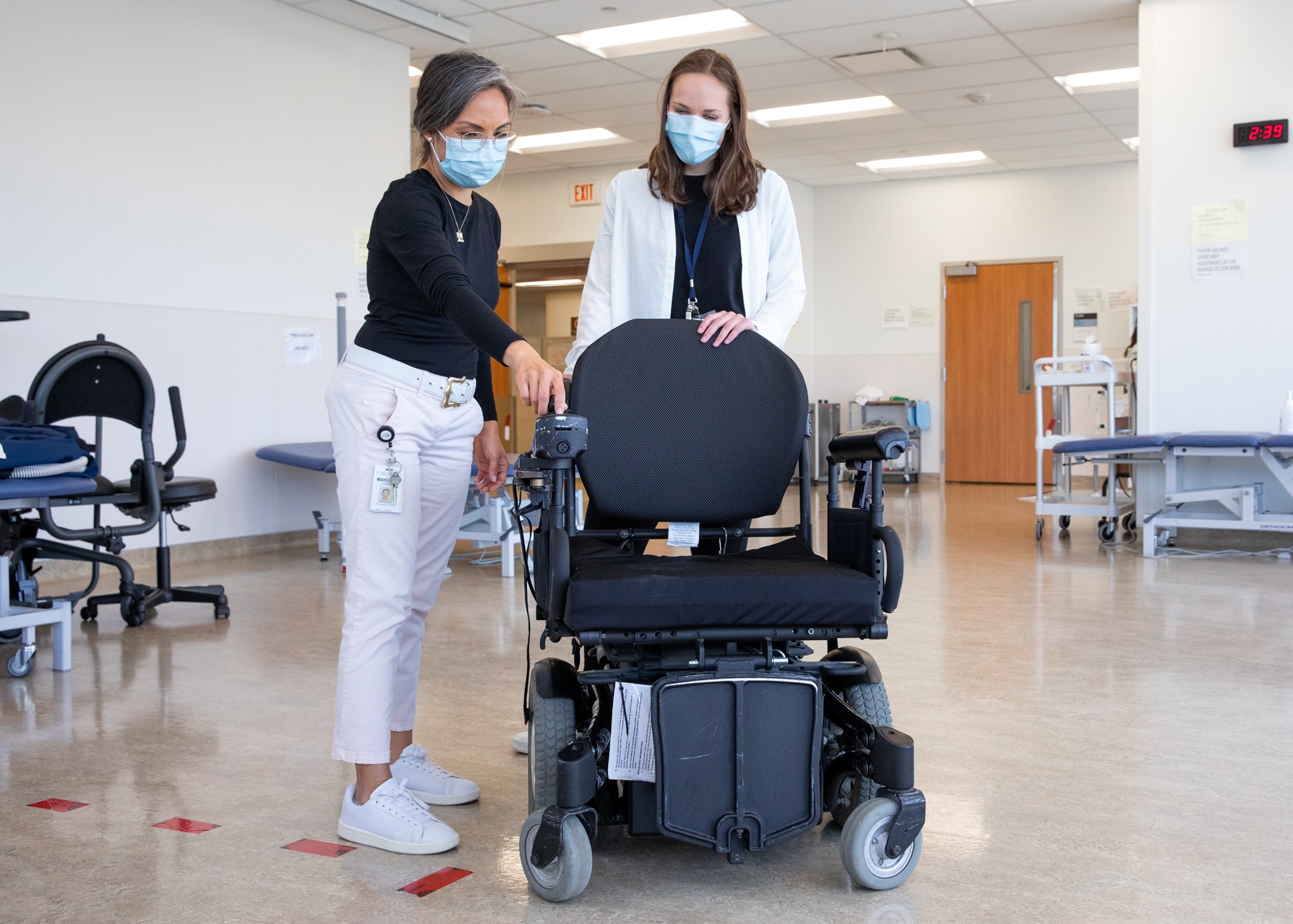 A Health care professional and a health care learner looking at a power wheelchair. The health care professional is demonstrating the controls on the armrest of the wheelchair.