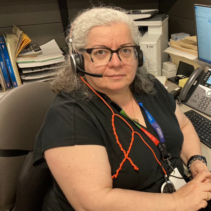 A woman is sitting at a desk looking at the camera, smiling. She is wearing a telephone headset and there is phone and computer keyboard behind her. 