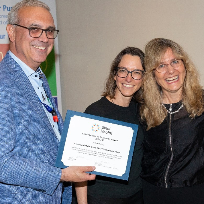 A group of three people one is holding a certificate to present to the other two people. They are looking at the camera smiling
