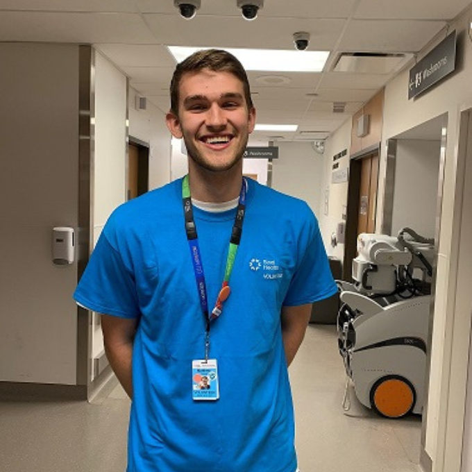 Portrait of a man standing in a hospital hallway wearing a turquoise volunteer t-shirt.