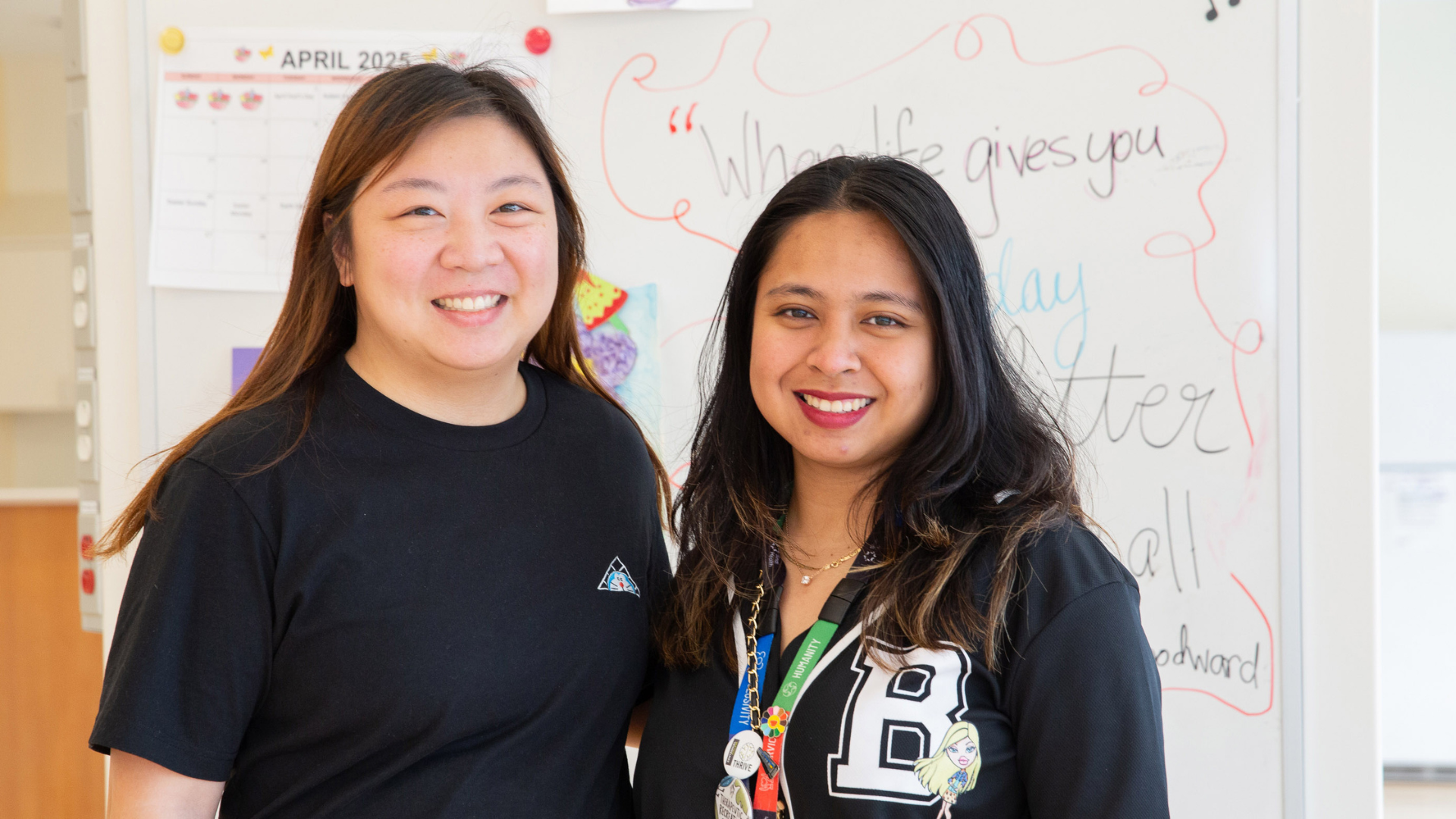Two women, standing looking at the camera smiling. They are standing in front of a white board with writing on it and a calendar