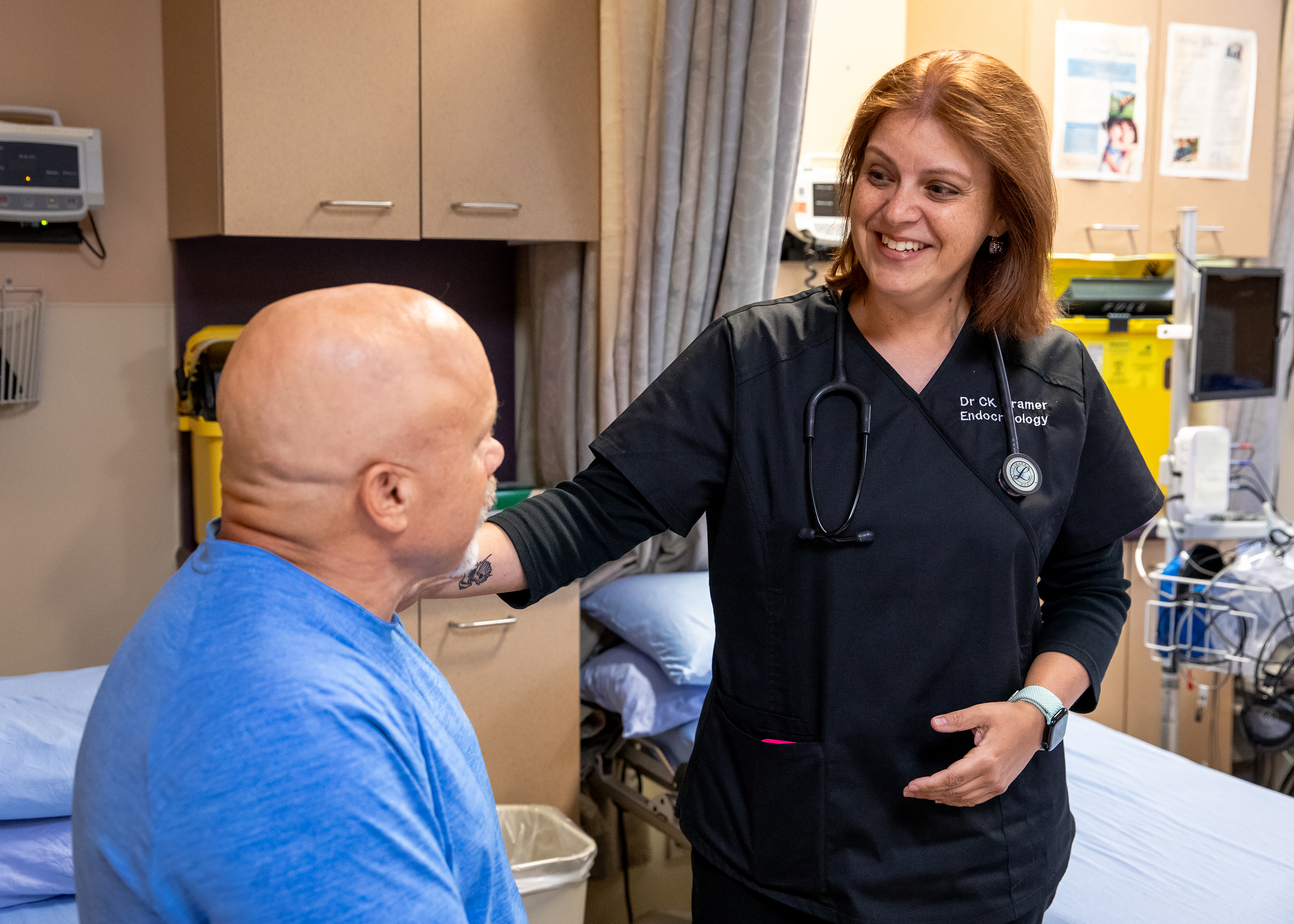 A smiling female doctor in black scrubs with a stethoscope labeled "Endocrinology" places a reassuring hand on the shoulder of the man, who is seated and facing her in a medical room.