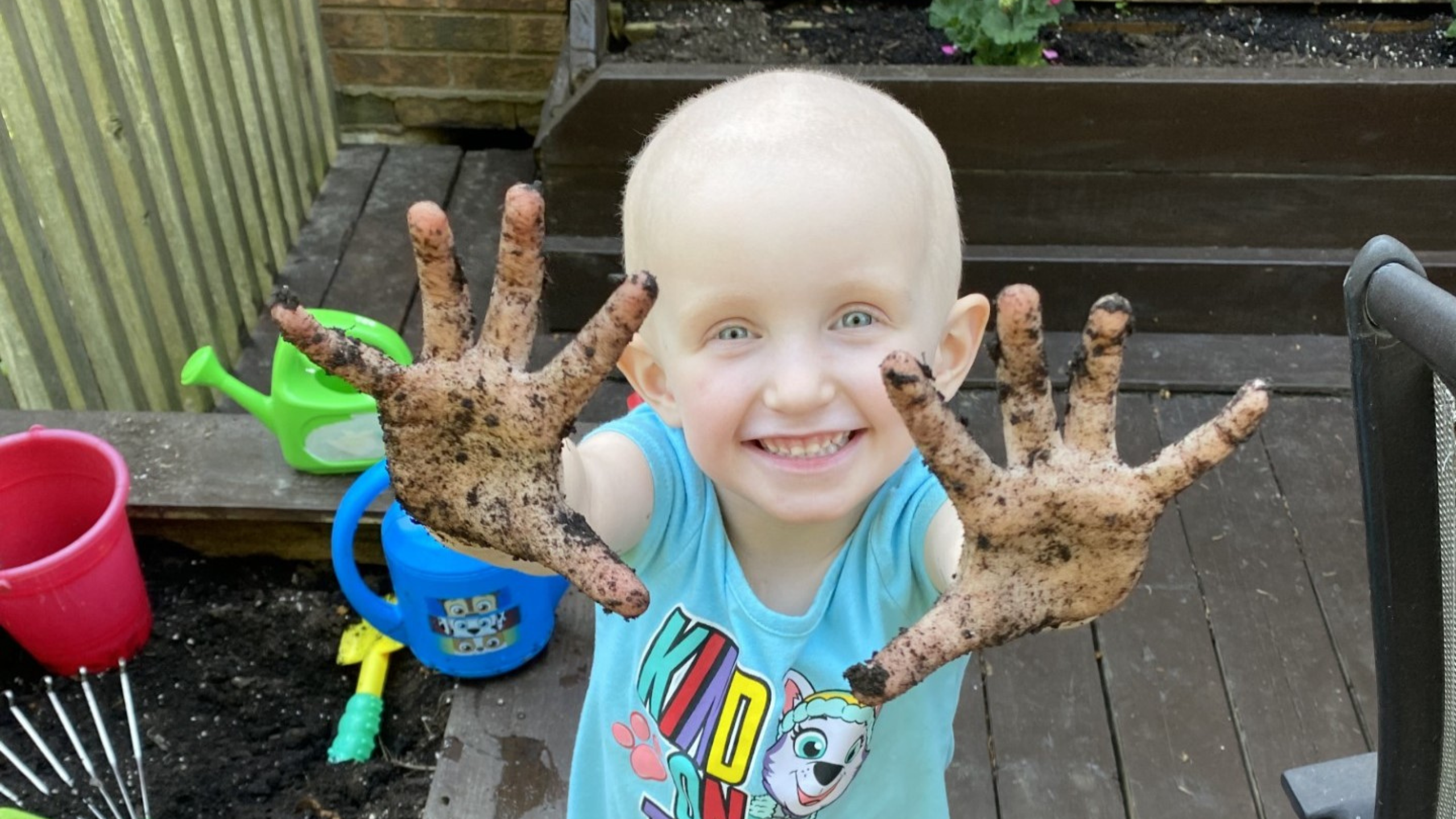 child holds up their hands, covered in dirt, with sandbox behind them