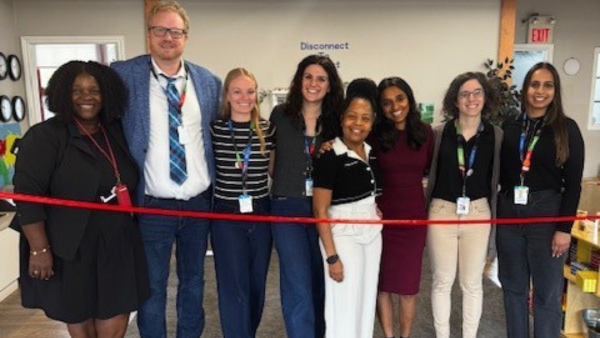 A group of people standing in a line, side-by-side. They are looking at the camera, smiling They are standing in front of a ribbon 