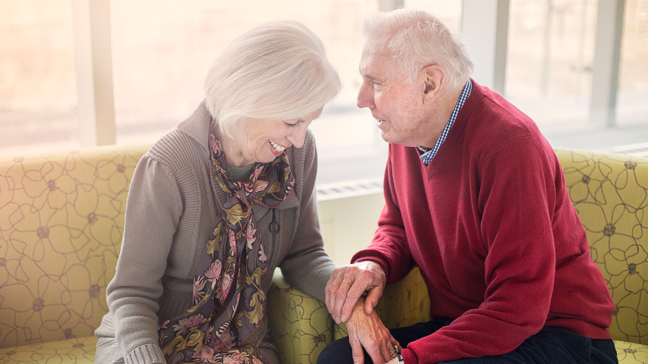 Woman and man both with grey hair sitting on yellow chairs holding hand with their faces close together