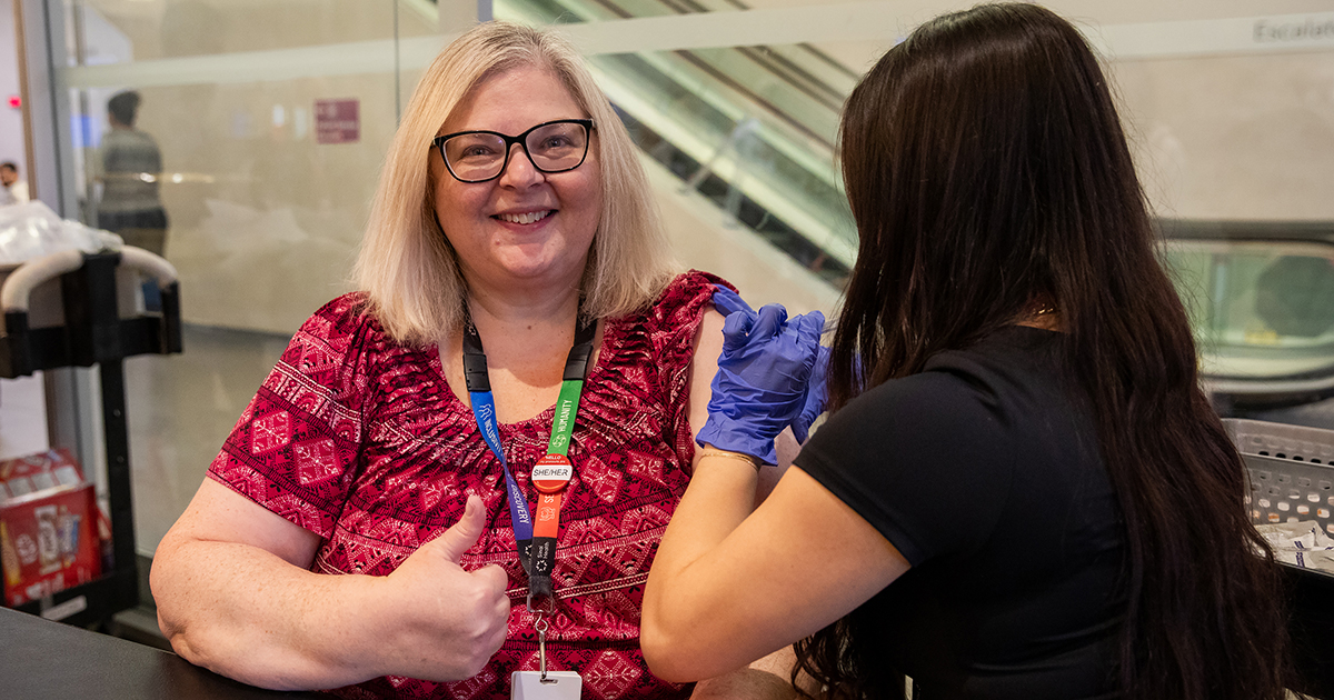 A woman gives the thumbs up while receiving a vaccination from a nurse. They are both sitting at a table.