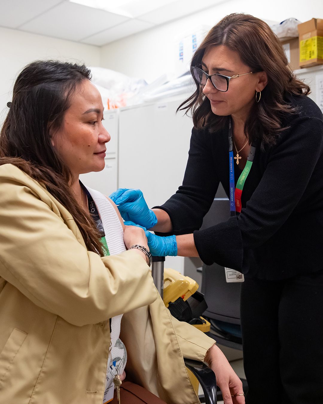 A woman receives a vaccination from a nurse. The woman receiving the vaccination is seated and the nurse is standing by leaning over her shoulder. they are in an office