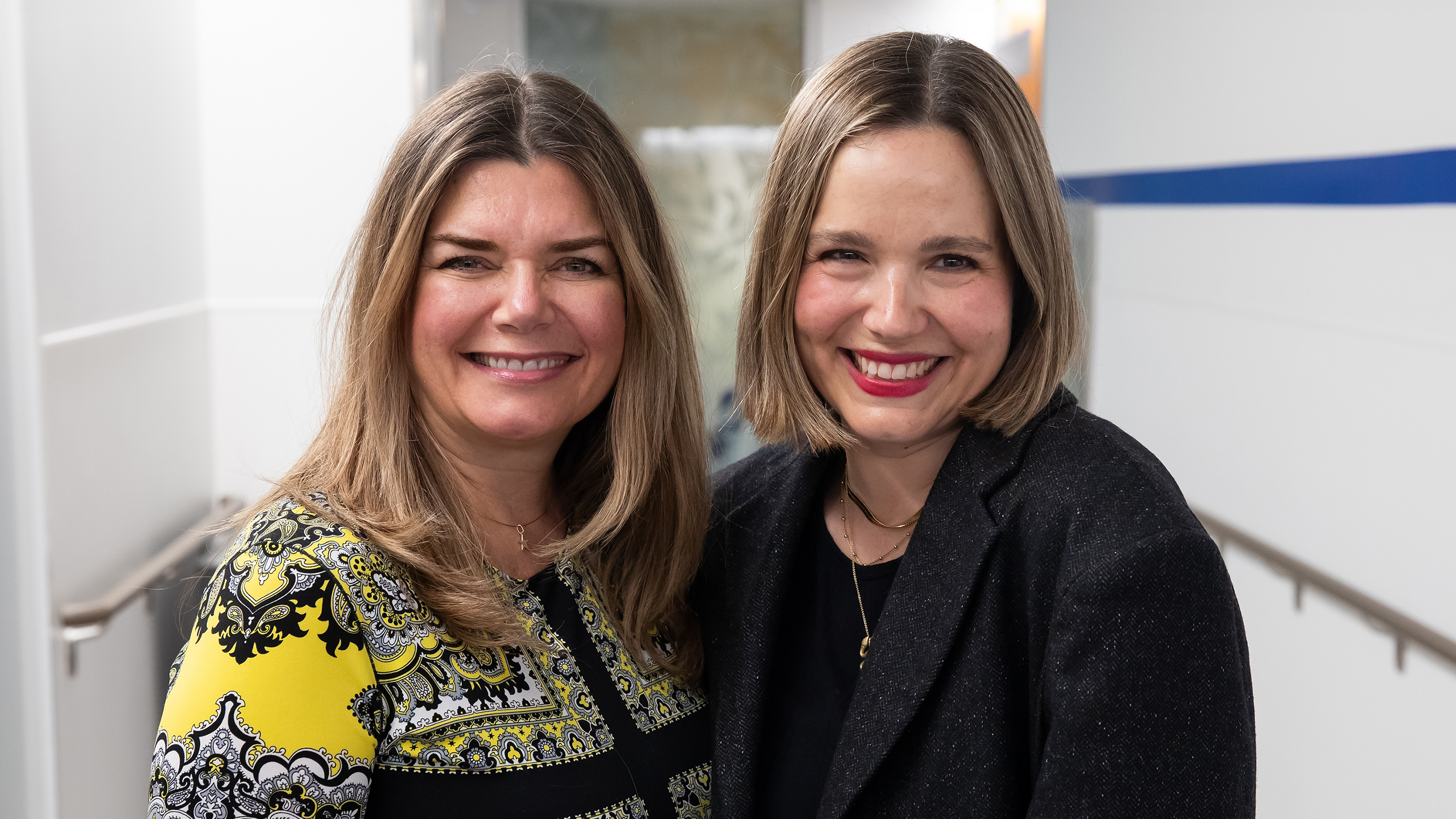 Two blonde women standing close together in a hallway smiling