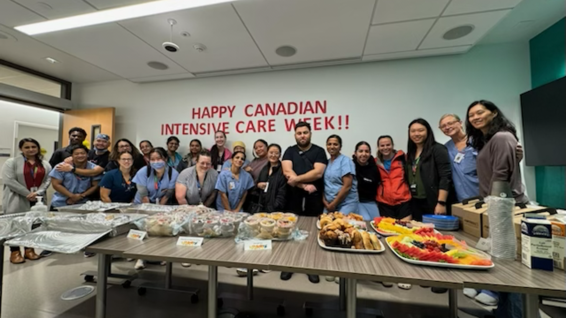 A large group of hospital employees standing in a group, looking at the camera smiling. There is a sign that says 'Happy Intensive Care Week," on the wall behind them and a table of food items in front of them.