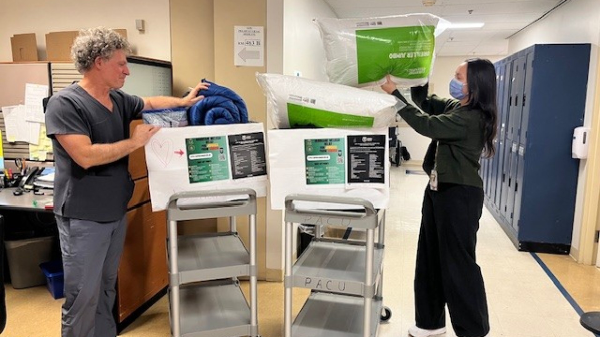 Two health-care workers put items including a pillow and bed roll in a box. They are in a corridor of a hospital