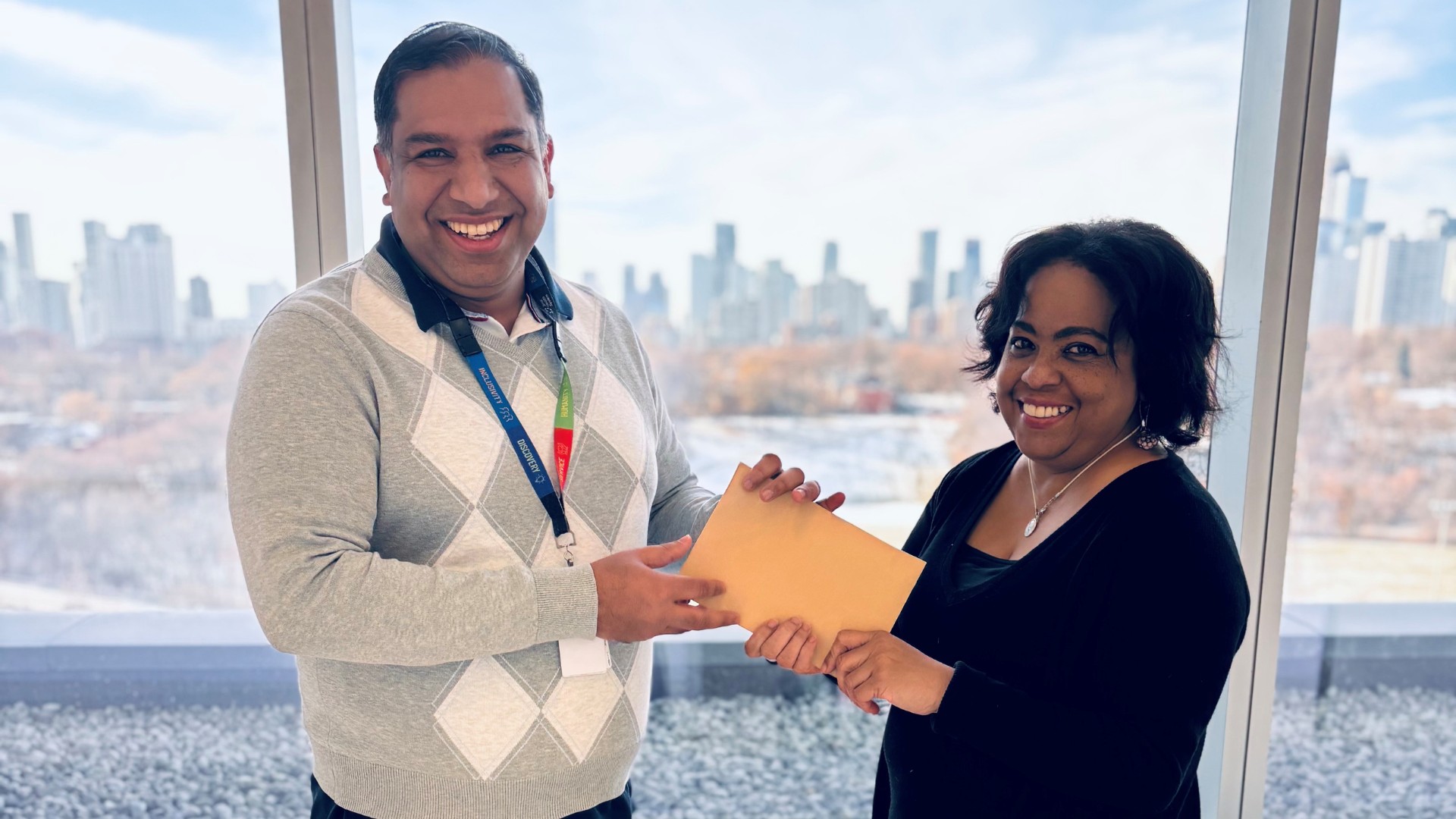 Two people stand in front of a window, one is handing an envelope to the other. They are looking at the camera, smiling