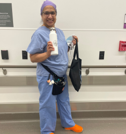 A health-care professional wearing scrubs stands in a hospital hallway. She is holding a reusable shopping bag and water bottle. Looking at the camera, smiling.