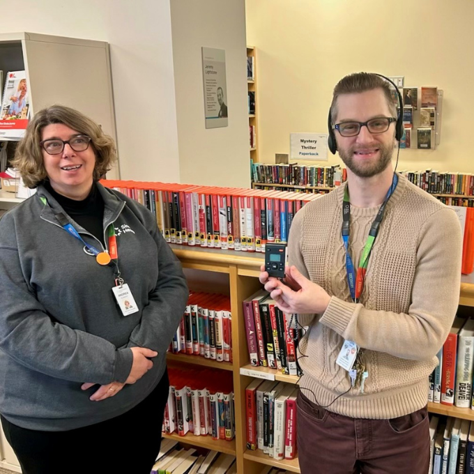 Two people standing side-by-side in a library. They are facing the camera and smiling. One is wearing headphones and holding up a small device that plays an audiobook.