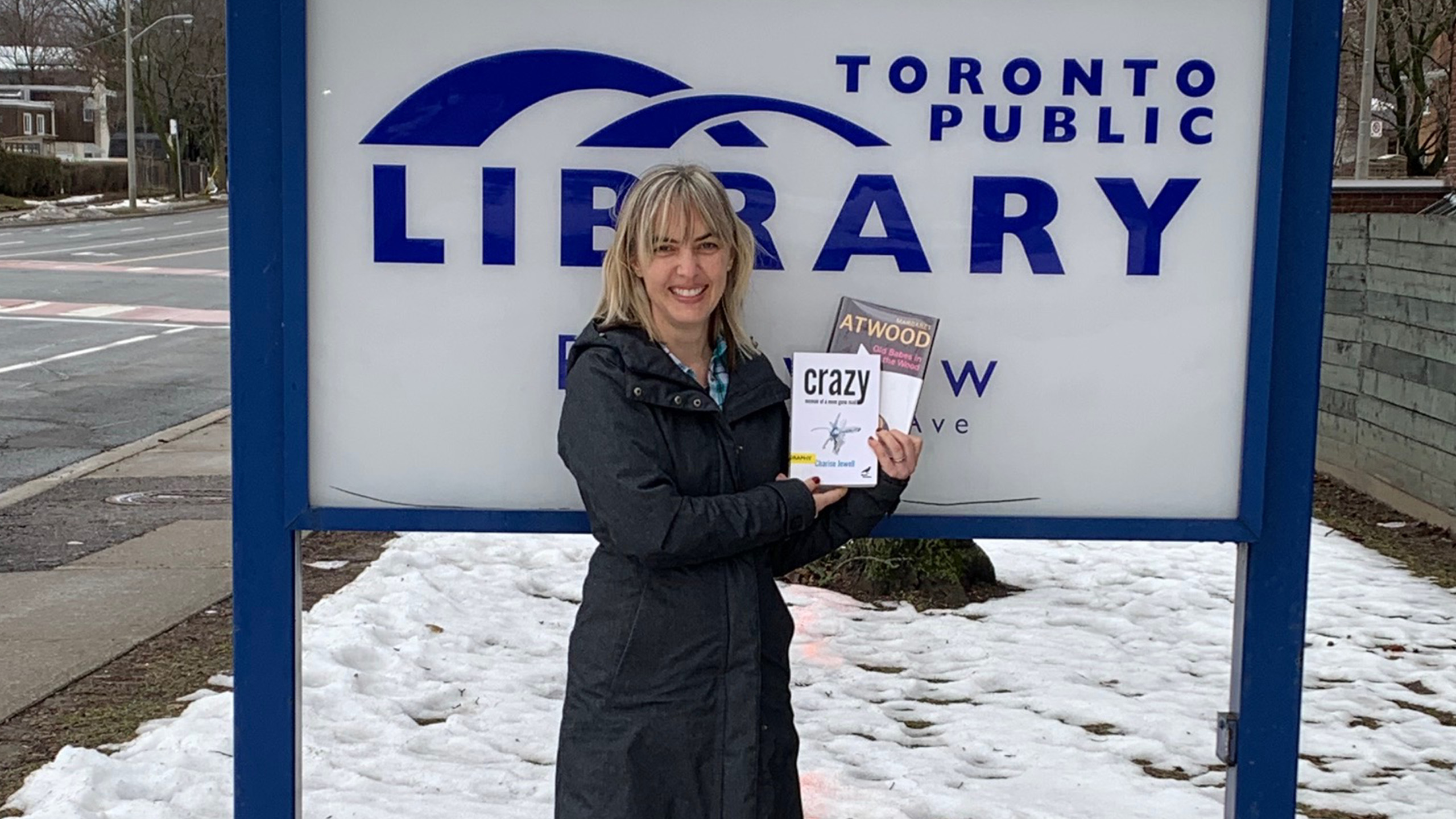 Woman stands in front of Toronto public library sign holding book