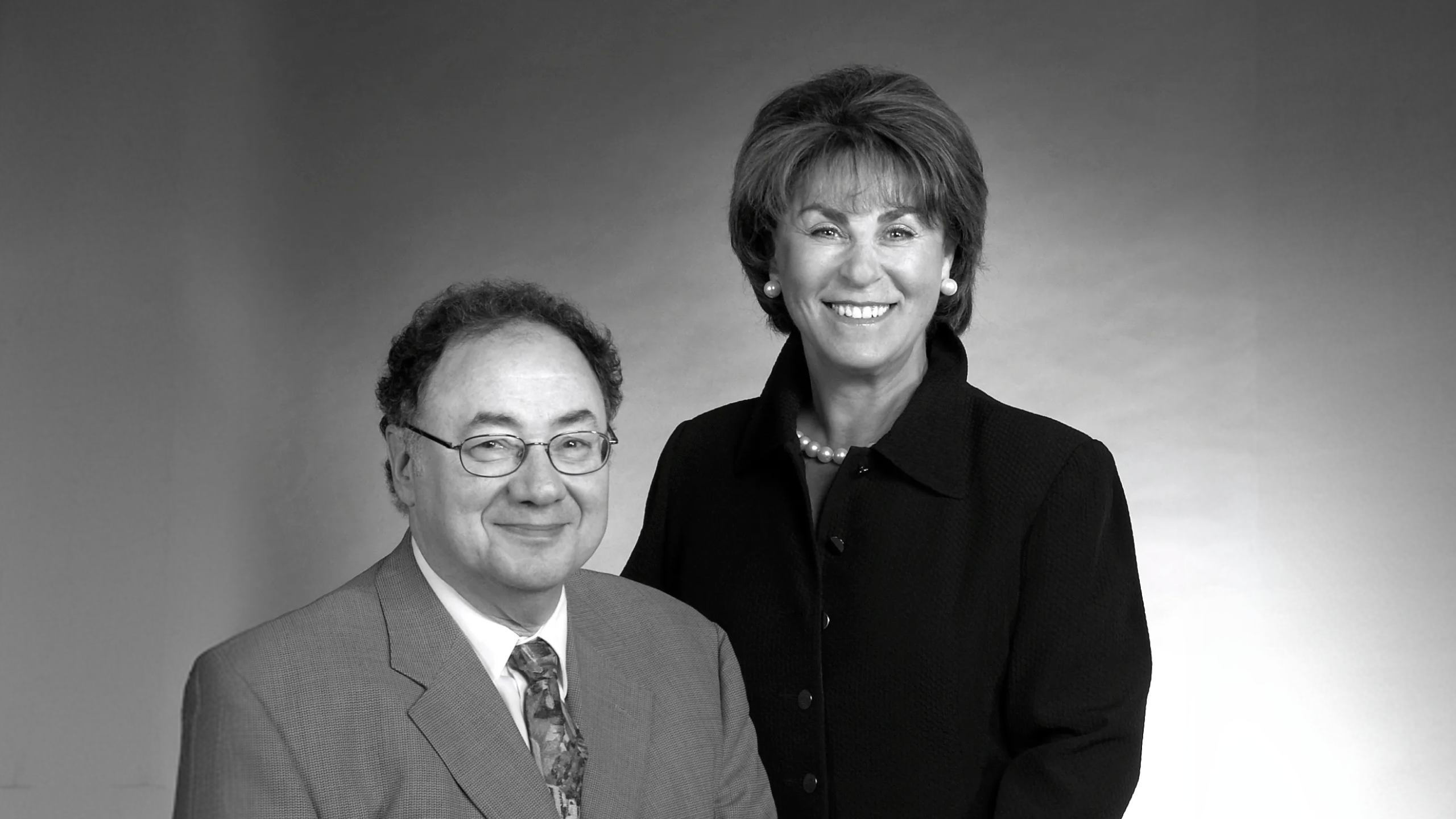 Black-and-white portrait of Barry Sherman seated in a suit and tie, with Honey Sherman standing behind in a dark jacket and pearls.