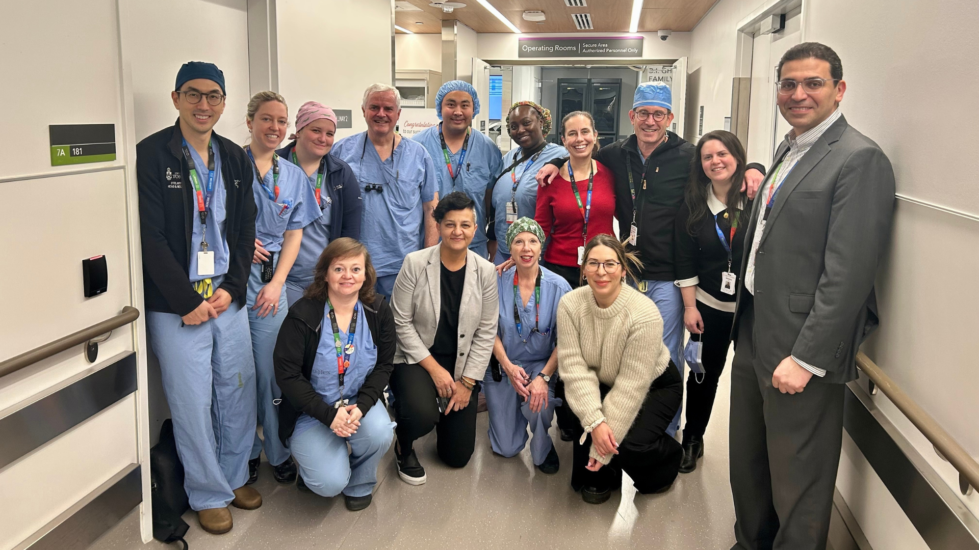 A group of health-care workers standing in a hospital in a hallway. Some are wearing surgical scrubs. They are facing the camera, smiling 
