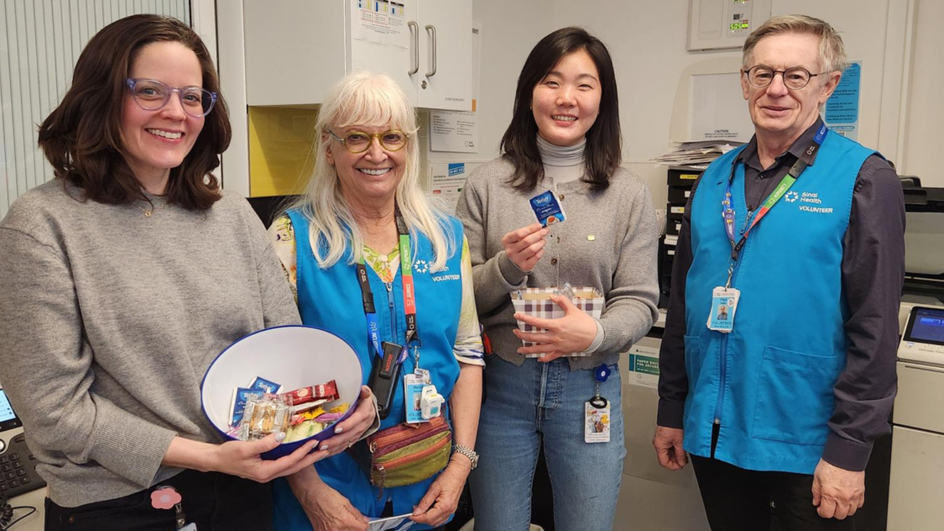 A group of four people standing in a hospital. Two are wearing vests of volunteers at Mount Sinai Hospital, the other two are employees. One is holding a large bowl that has some packaged snacks. They are standing and looking at the camera smiling