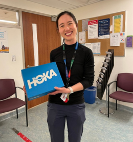 A Sinai Health employee stands in a waiting room. She is holding up a shoe box and looking at the camera smiling.