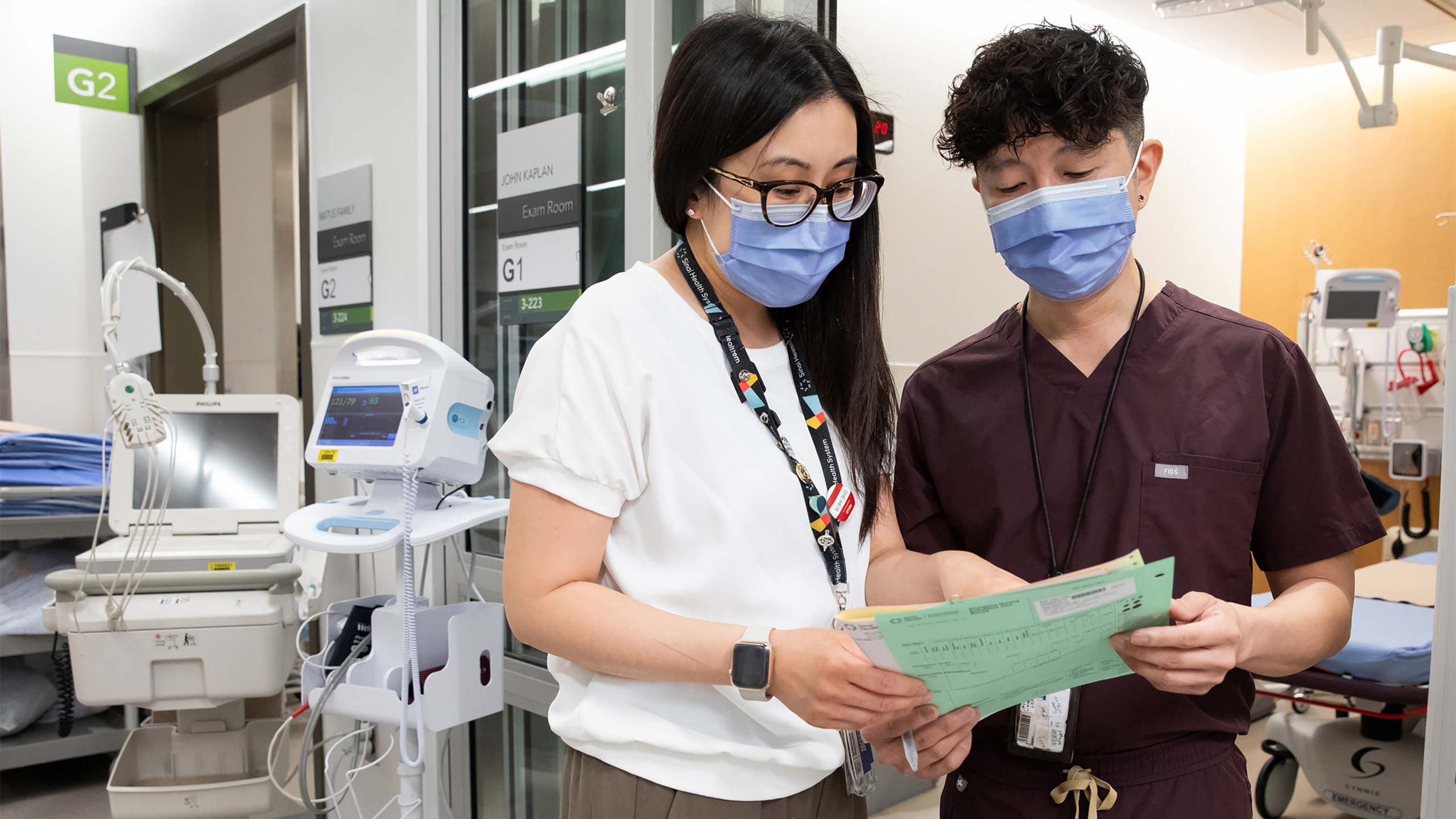Woman and man both wearing medical masks. They standing are looking at a piece of paper with medical equipment in the background behind them.
