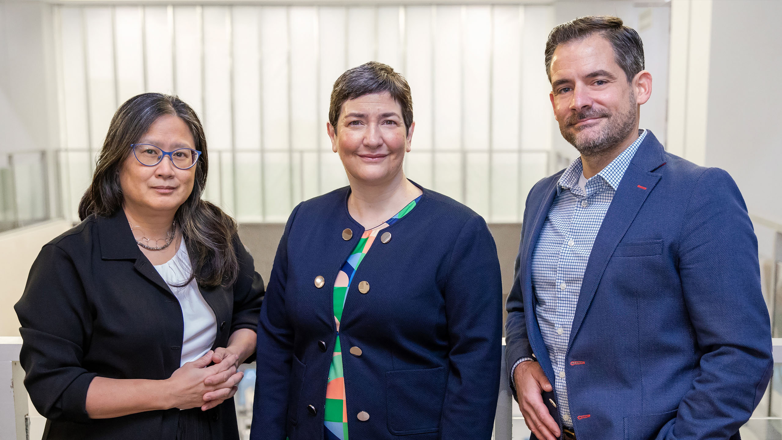 A women in a black blazer, a woman in a blue blazer and a man in a blue blazer stand together leaning against a railing with a bright background behind them