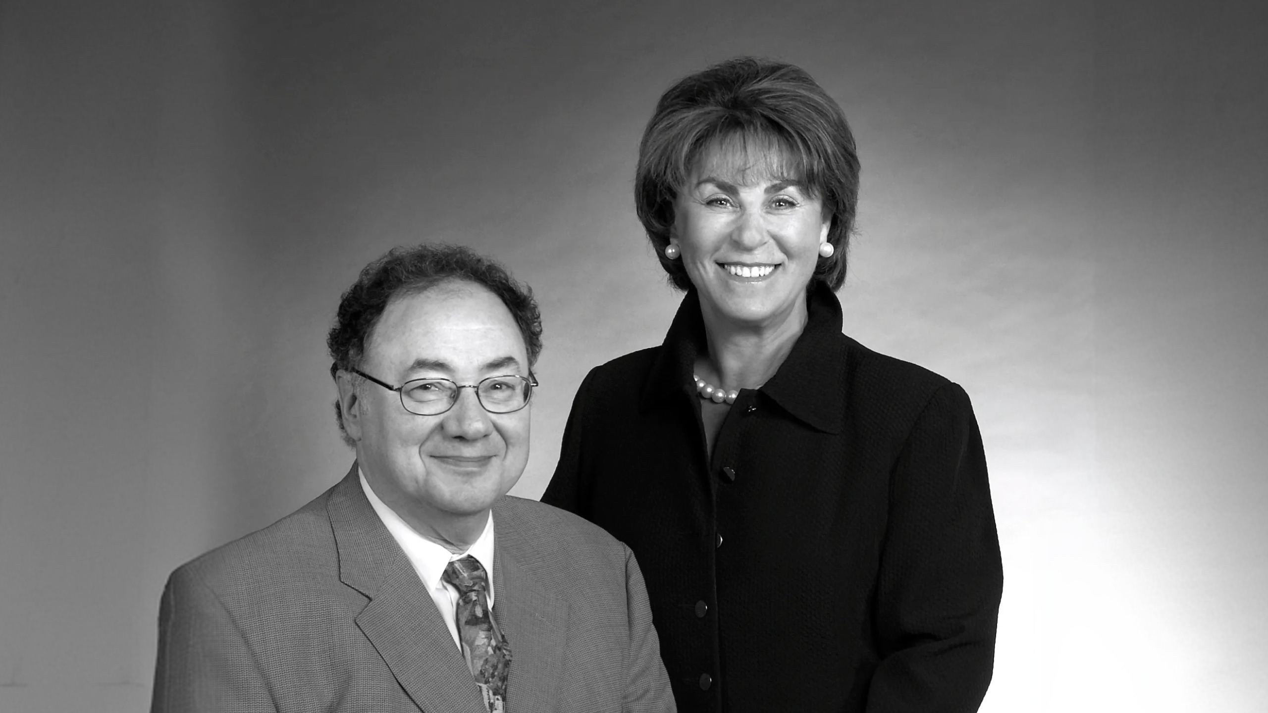 Black-and-white portrait of Barry Sherman seated in a suit and tie, with Honey Sherman standing behind in a dark jacket and pearls.