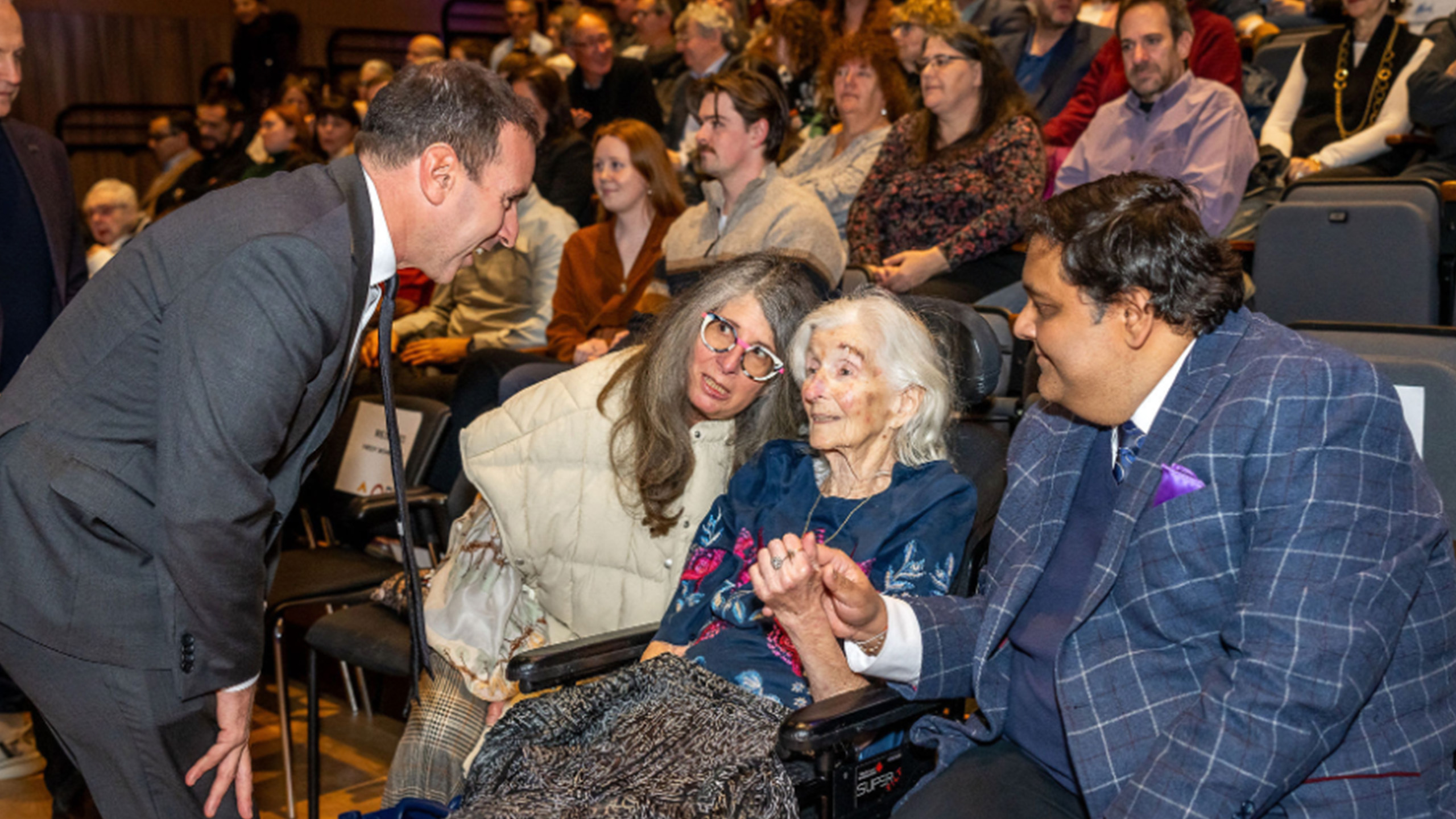 man in suit speaks to an elderly woman sitting next to woman and man