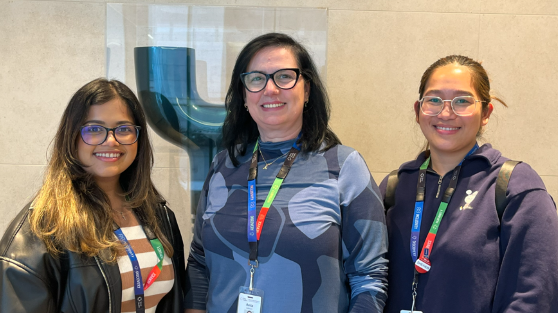 three women standing side-by-side. they are looking at the camera, smiling. There is a beige wall in the background