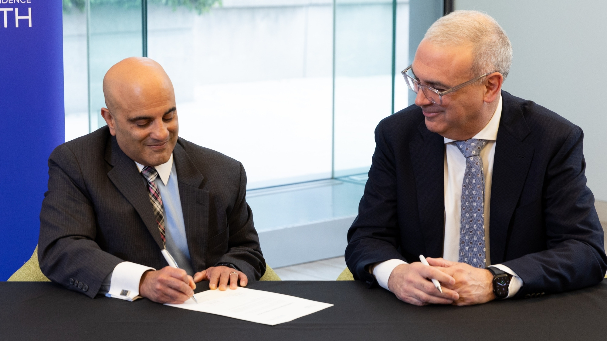 Dr. Newton and Altaf Stationwala sitting at a table signing a symbolic paper to celebrate the partnership.