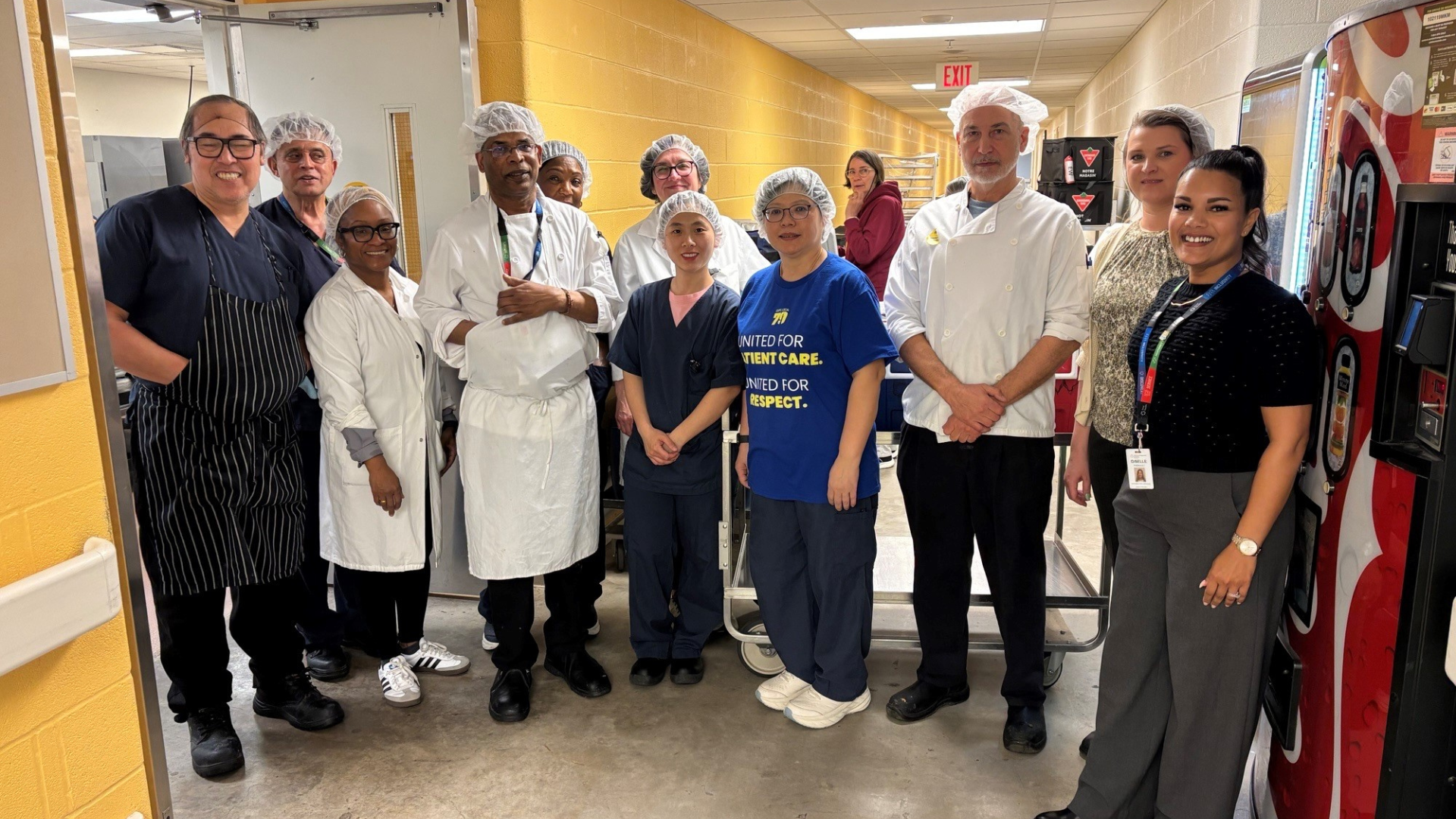 A group of people who work in Food Services at Hennick Bridgepoint Hospital stand in a hallway just outside the kitchen. Some are wearing hairnets and scrubs. They are looking at the camera, smiling