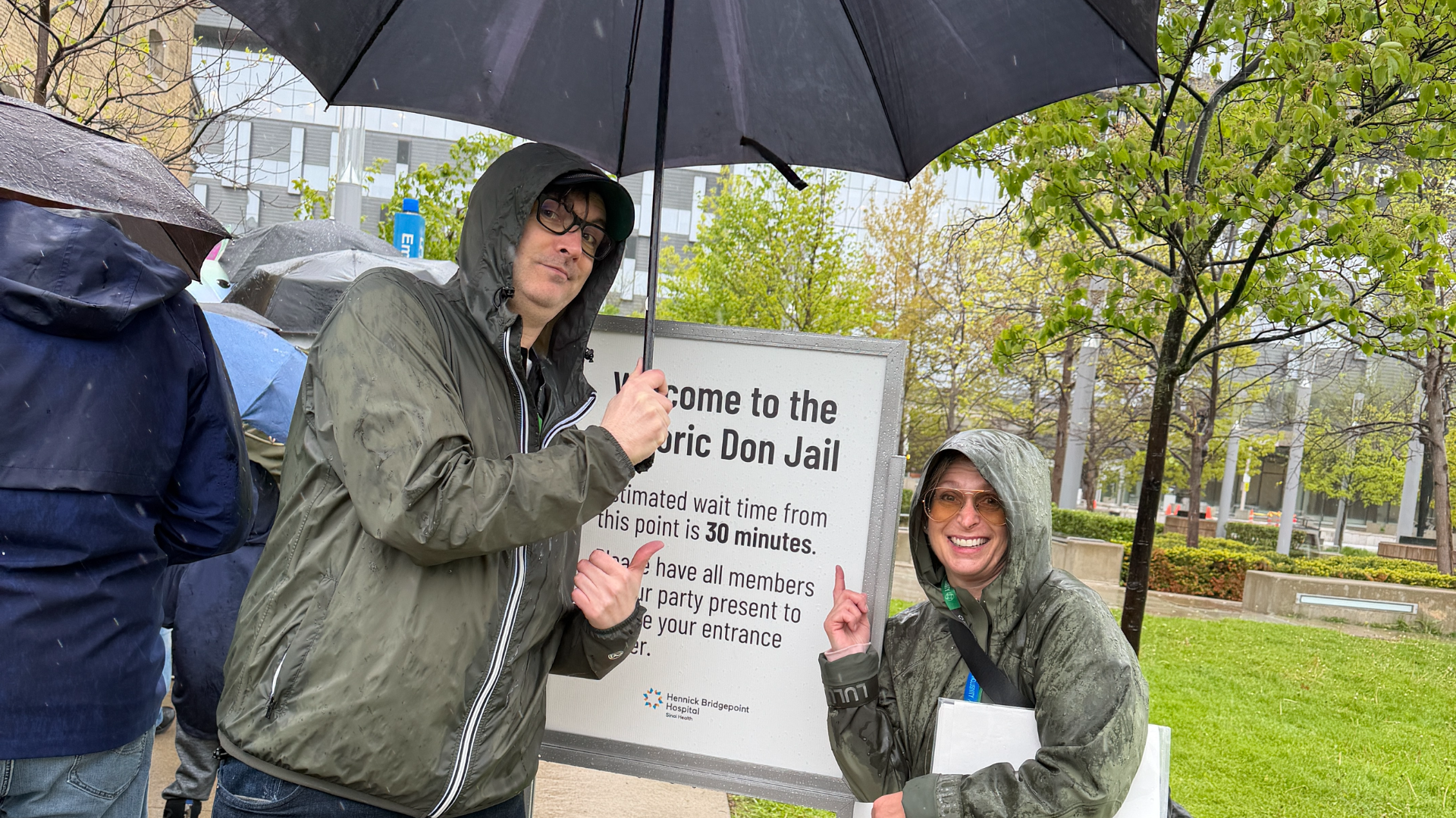 Two Sinai Health employees wearing rain-coats, stand outside in front of a sign for the Doors Open event. 