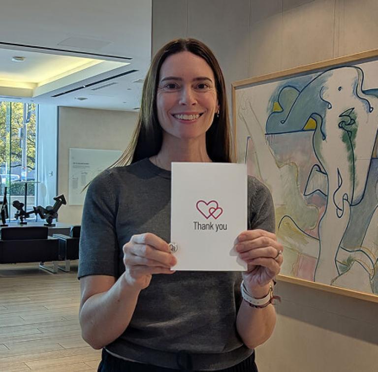 A woman in a lobby holds a “Thank you” card and smiles at the camera.