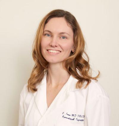 Medium close shot of a woman with shoulder length blond hair, wearing a white lab coat and smiling. 