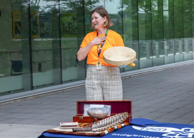 A woman in an orange shirt holds an indigenous ceremonial drum. Materials used for a smuding ceremony sit on a table in front of her
