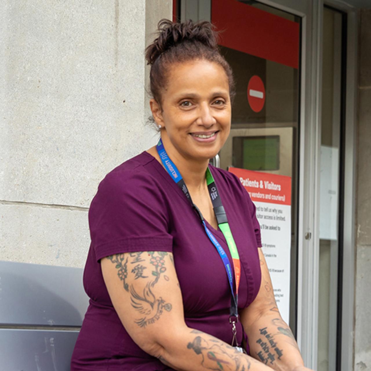 A health care worker wearing scrubs and a lanyard with an identification badge stands outside a hospital looking at the camera smiling