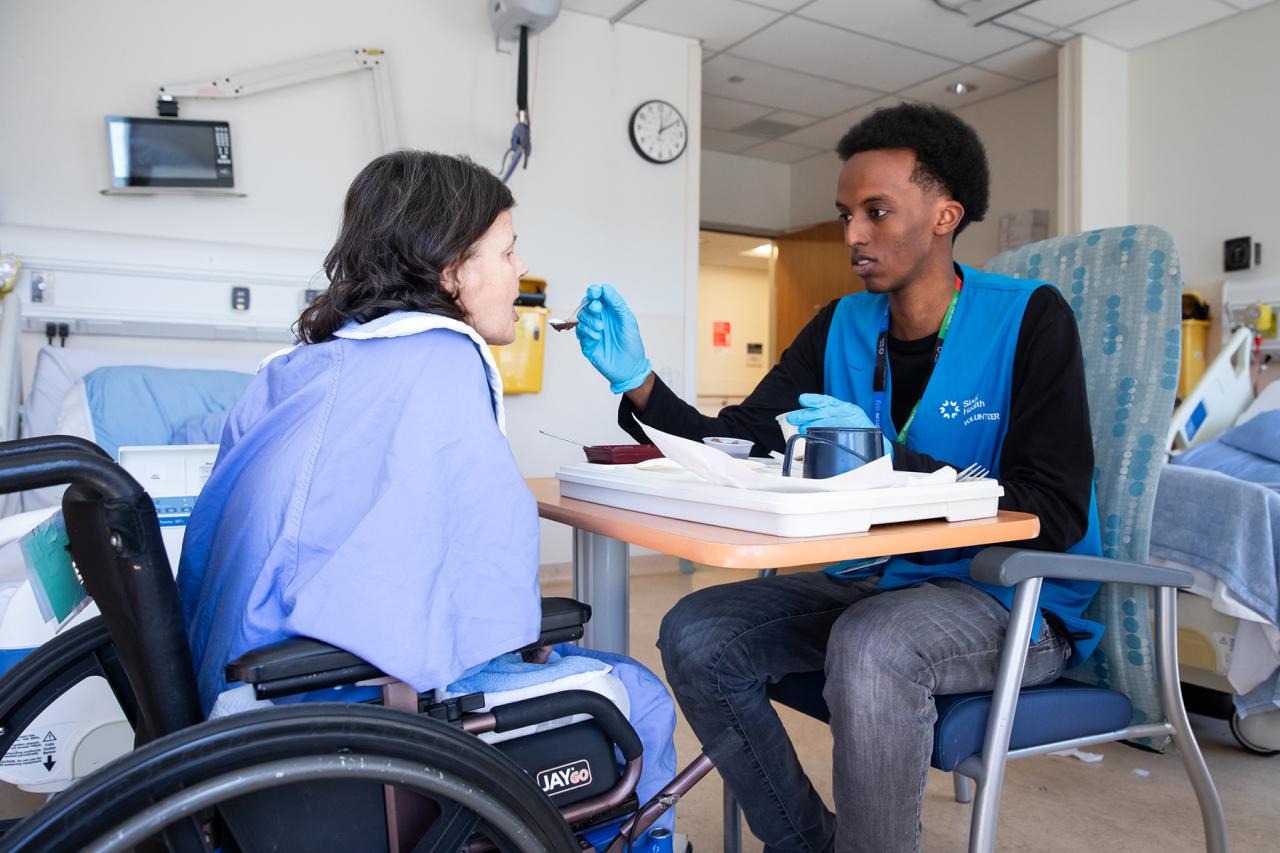 A hospital volunteer lifts a spoon to support a patient with a meal.