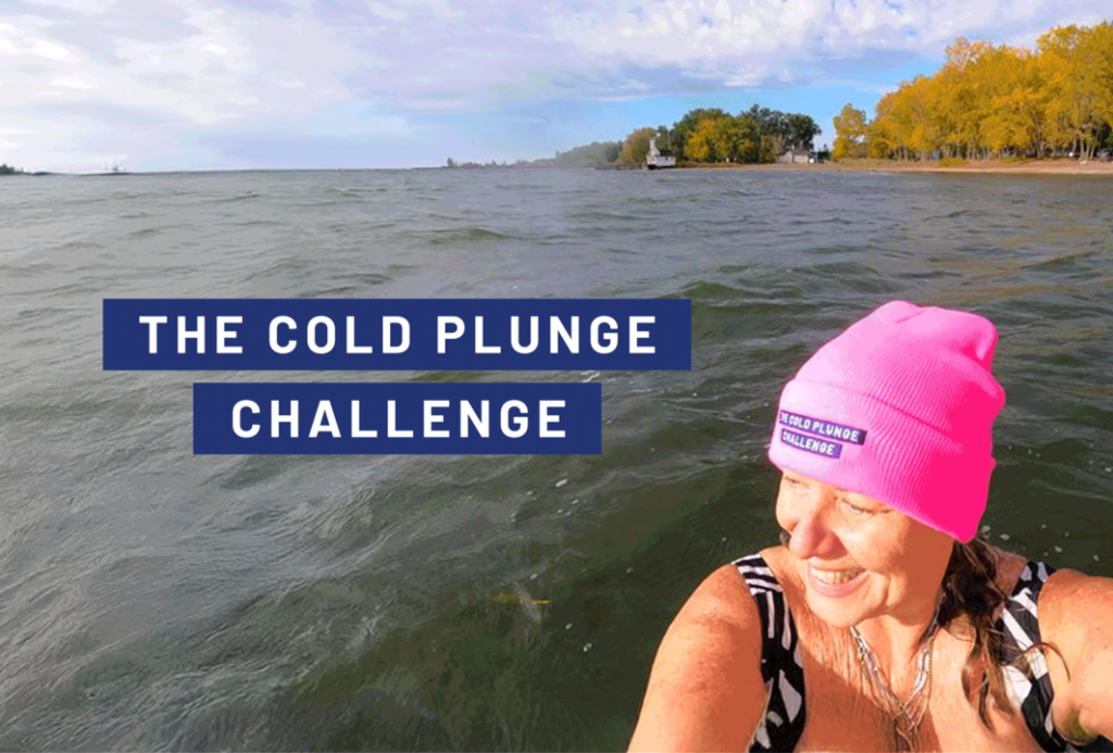 A person wearing a bright pink winter hat stands in cold lake water near a shoreline lined with autumn trees. Another participant is visible farther out on the right. Text over the water reads “The Cold Plunge Challenge” against a blue background.