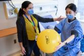 A physiotherapist looks at a patient who sits on a hospital bed and holds a yellow exercise ball