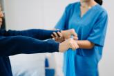 A health-care provider assists a patient as he lifts his arms in front of him while holding a blue exercise band