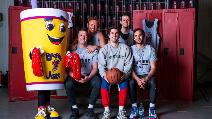 Five basketball teammates arranged in two rows in front of red lockers, smiling next to the Booster Juice mascot. Three sit in the front with the middle player holding a basketball, and two stand behind