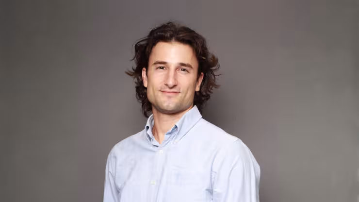 Tal Schwartz, smiling, wearing a blue button-up shirt against a light grey backdrop