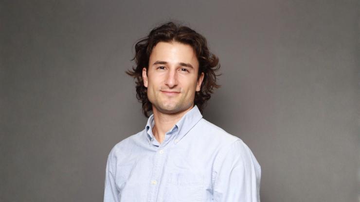 Tal Schwartz, smiling, wearing a blue button-up shirt against a light grey backdrop