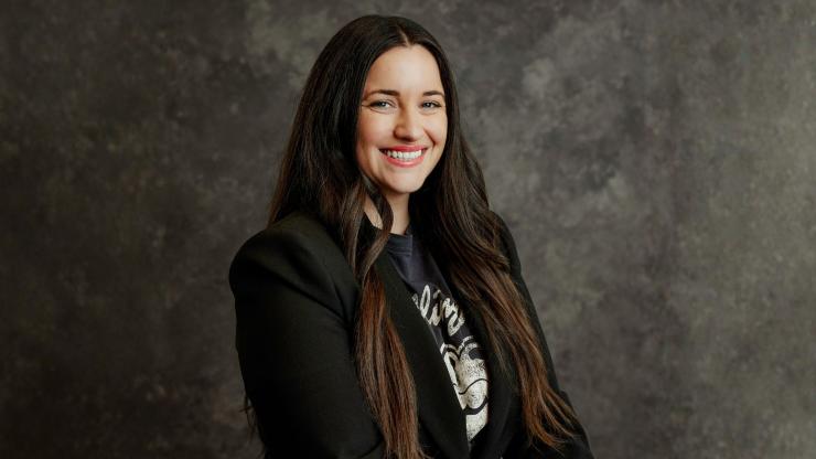 Erin Sawala, smiling, wearing a black blazer against a dark grey backdrop