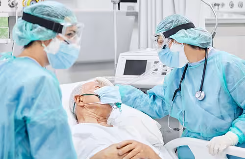 Two health care providers, wearing blue scrubs stand with a patient in the ICU 