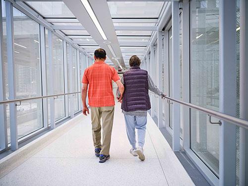Medium long shot a man, wearing an orange checked shirt and khaki pants, holds hands with a woman, wearing a purple vest and grey sweat pants. They are walking down a hallway with windows with their backs to the camera.