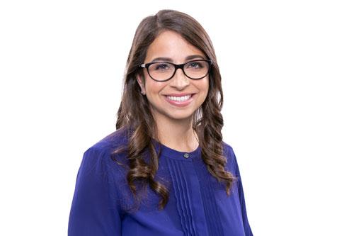 Medium close shot of a smiling woman with long, curled brown hair, wearing a purple blouse and black glasses