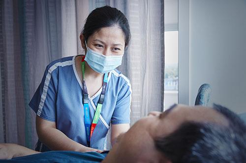 An Asian nurse wearing blue scrubs, a mask, and a colourful lanyard leans towards a man who is laying down.