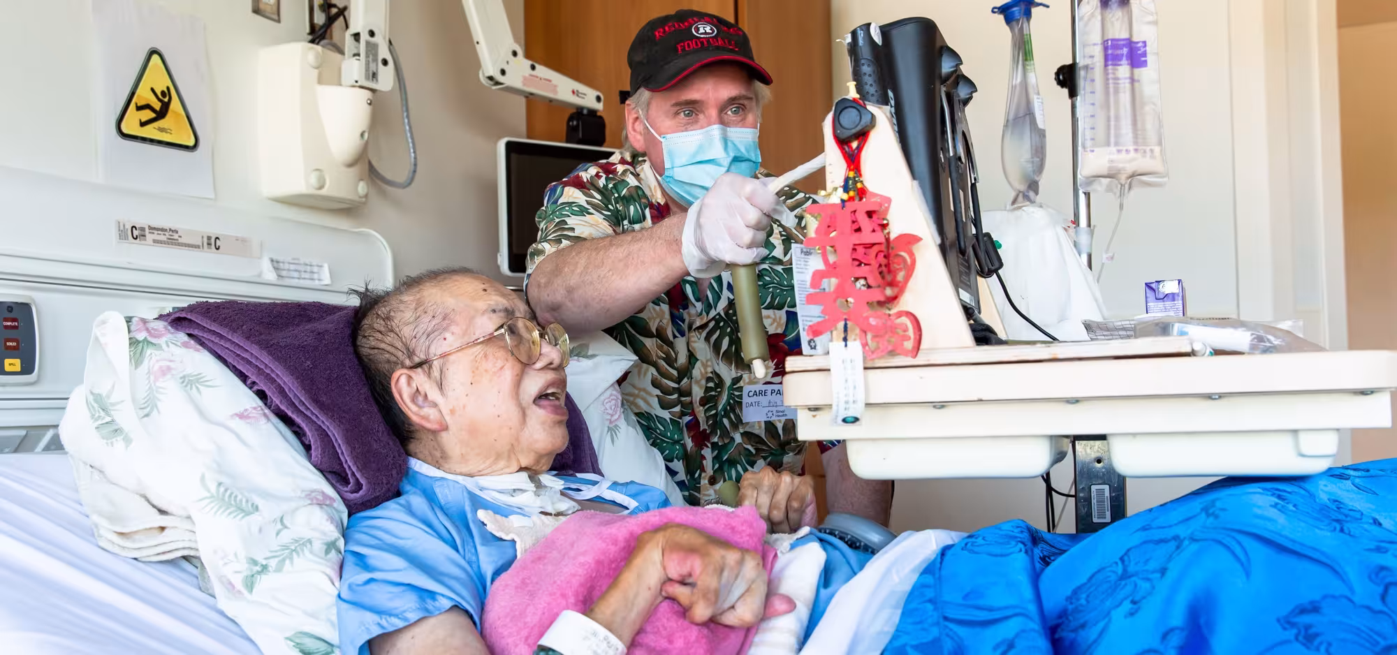 A woman in a hospital bed with her husband standing next to her. They are both looking at a computer screen and he is tapping the screen with a stylus