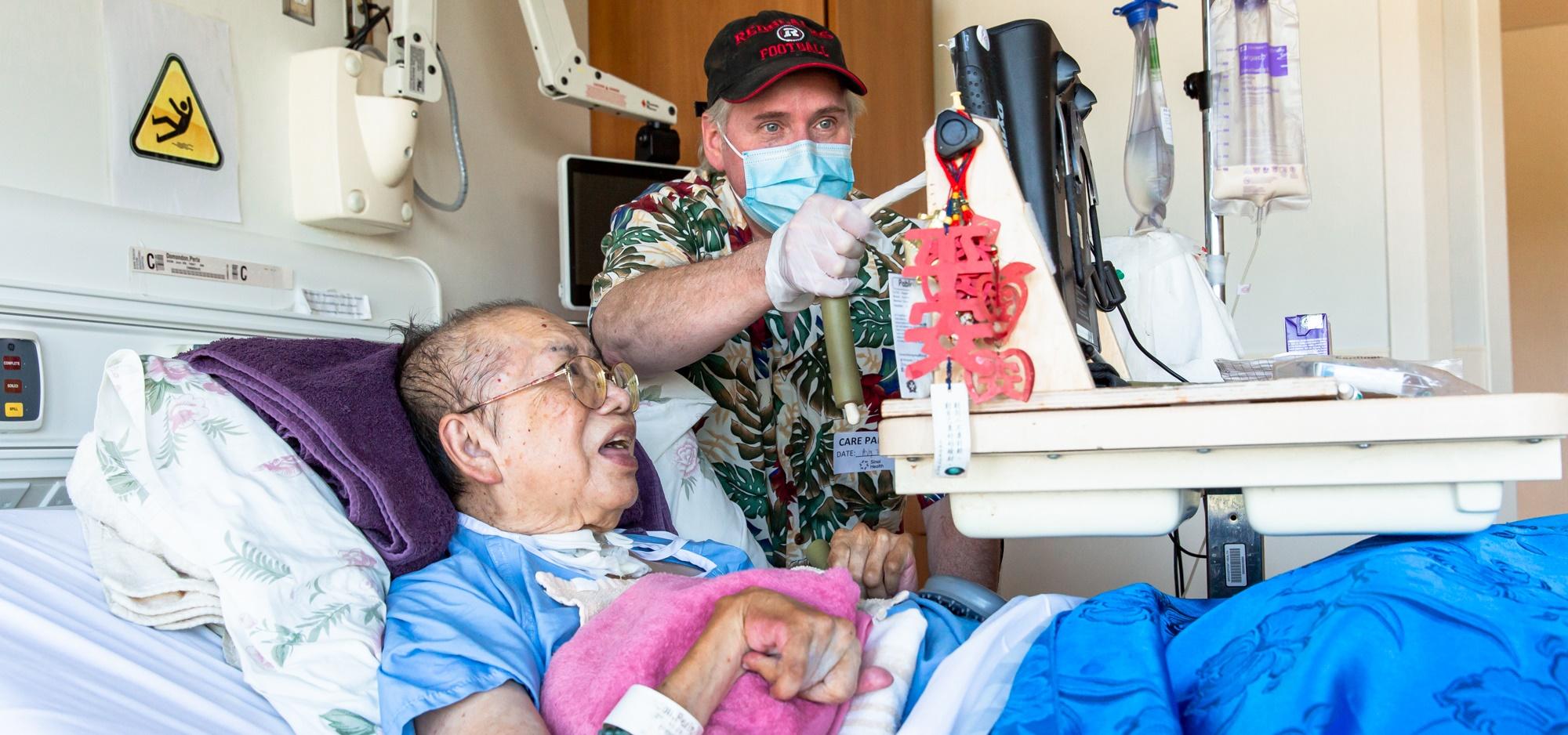 A woman in a hospital bed with her husband standing next to her. They are both looking at a computer screen and he is tapping the screen with a stylus