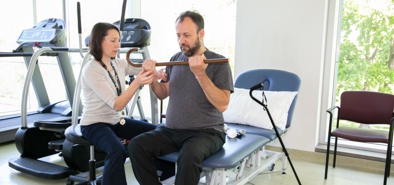 Physician helping man lift cane with both hands at rehabilitation centre.