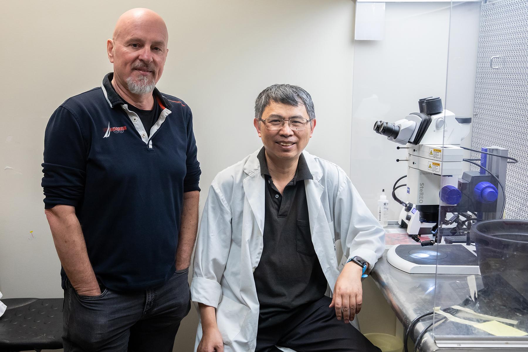 Two researchers pictured in the laboratory next to a microscope. One researcher. A Caucasian man is standing with hands in pockets and the other, a bespectacled Asian man, is seated and wears a lab coat.